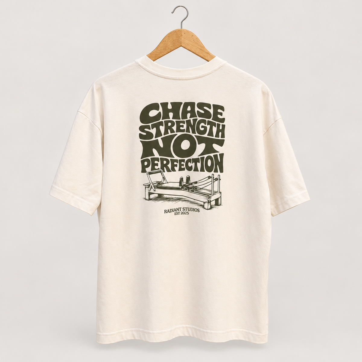 Cream Radiant Pilates T-shirt on wooden hanger, showing olive Chase Strength Not Perfection back graphic